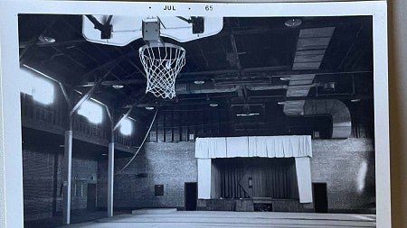 Photo from the 1960s shows Highland Hall when it was a gymnasium. A basketball hoop is at one end and a small stage at the other. 