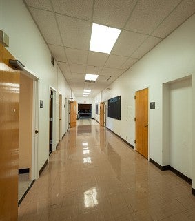 Before photo of a hallway in the Innovation Building. There are several light wood stained doors on each side, florescent lighting and tan linoleum floors.